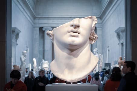 Parte inferiore di una statua in marmo esposta in un museo. Rappresenta una testa stile greco, divisa a metà in diagonale all'altezza del naso.